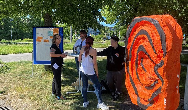 Vier Leute sprechen an einem Stehtisch neben dem orangenen plastischen Ohr 