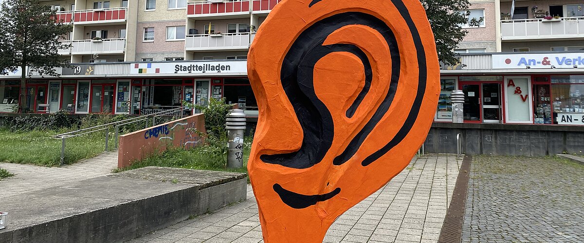 Ein großes orangenes plastisches Ohr steht vor einem Plattenbau.