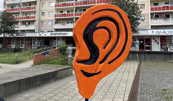 Ein großes orangenes plastisches Ohr steht vor einem Plattenbau.