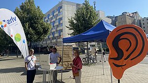 Drei Mitarbeitende der Volkshochschule stehen auf einem Marktplatz mit dem großen orangenen plastischen Ohr an einem Stehtisch.