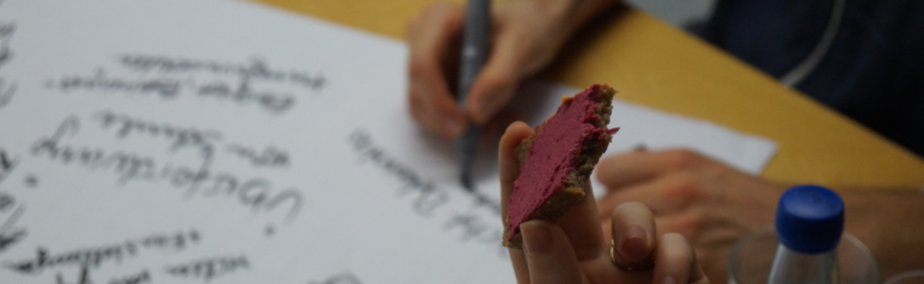 im Hintergrund wird auf ein flipchart geschrieben. im Vordergrund sieht man eine Hand mit einem belegten Brot.